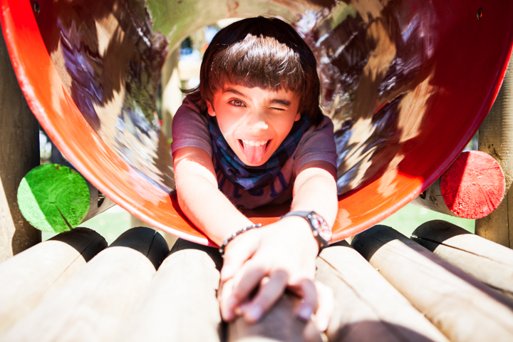 A laughing child reaches toward an adult from inside a colorful playground tunnel.