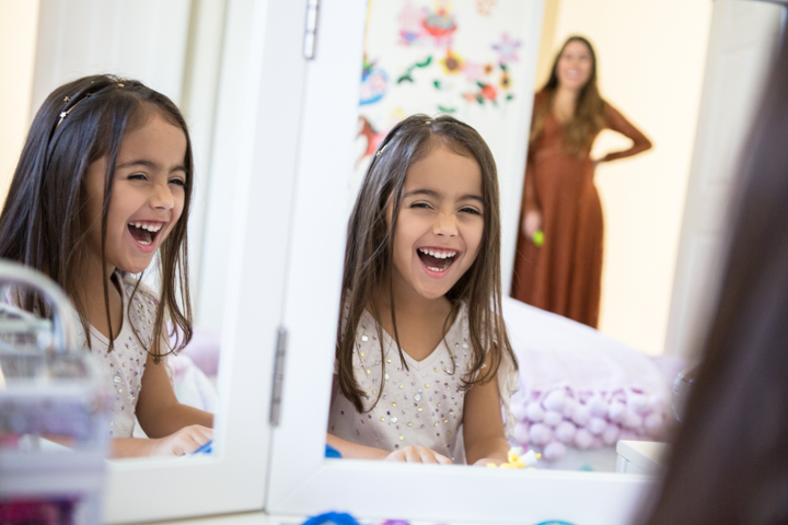 A laughing child looks into a mirror while a woman watches from a doorway.