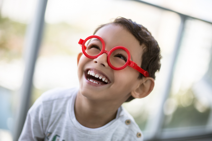 A laughing child wears round red glasses indoors.