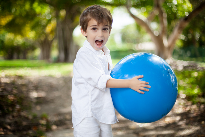 A child holds a large blue ball in a shady garden.