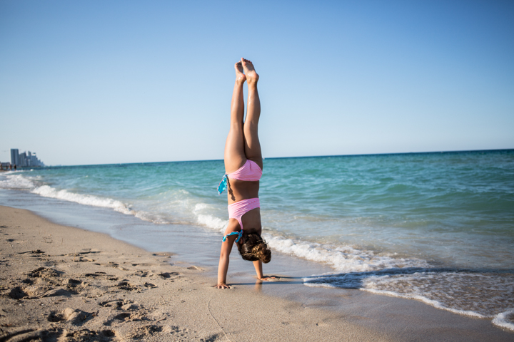 A child balances in a handstand on the sand beside the ocean.