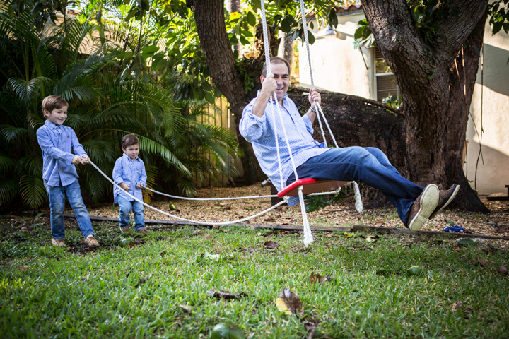 Two children pull a rope while a man sits on a garden swing.