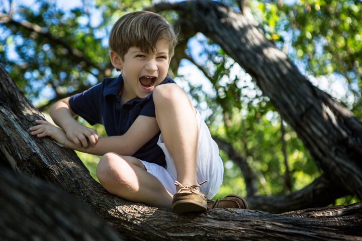 A laughing child crouches on a tree branch beneath green leaves.