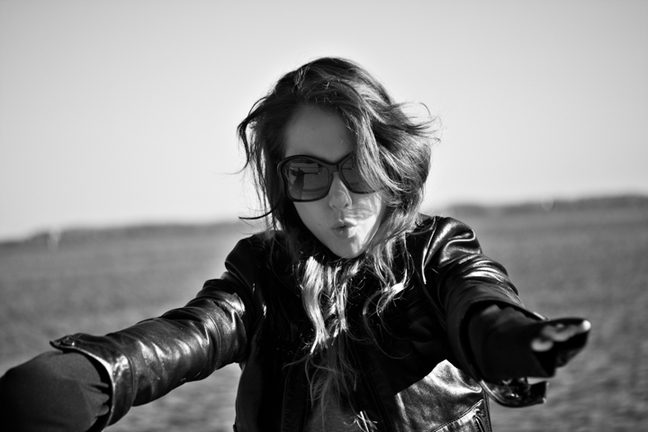 A woman in sunglasses and a leather jacket reaches toward the camera.