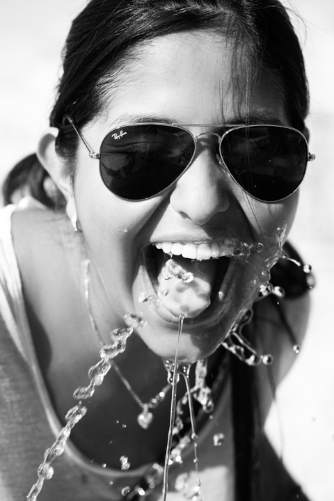 A person wearing sunglasses smiles with water splashing beside their open mouth in black and white.