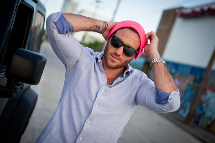 A man in a pink cap and sunglasses stretches beside a vehicle.