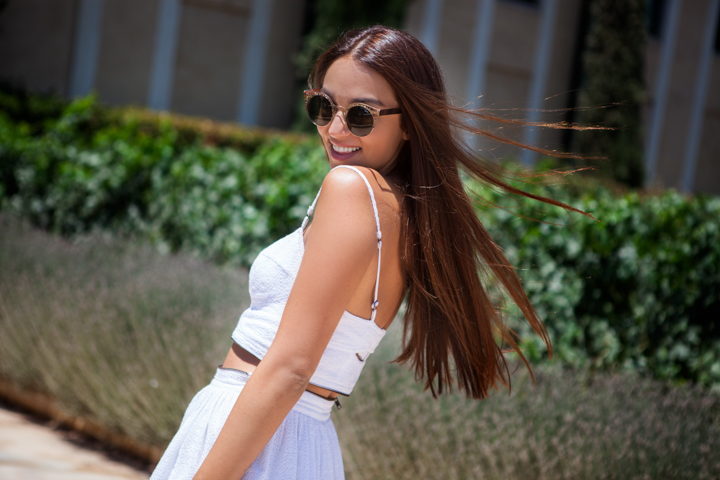 A woman in a white outfit turns toward the camera on a sidewalk.