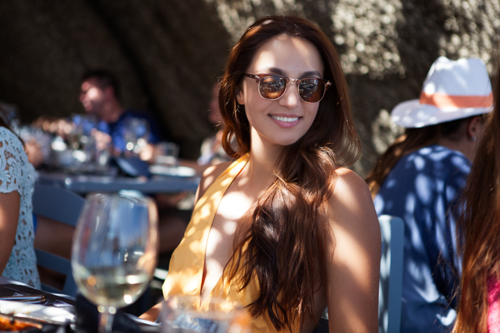 A woman in sunglasses turns toward the camera at an outdoor restaurant.