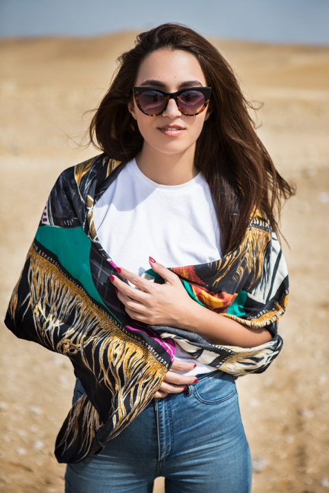 A woman in sunglasses and a patterned jacket stands in a dry landscape.