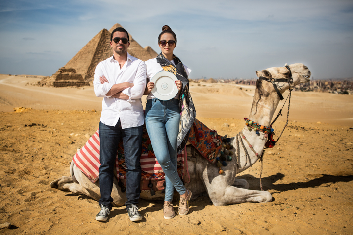 Two women stand beside a seated camel in a sandy landscape.