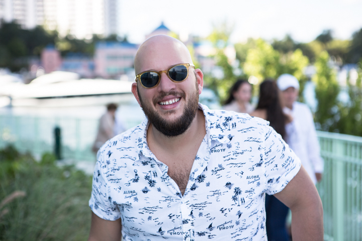 A smiling man in sunglasses and a patterned shirt near a waterfront railing.