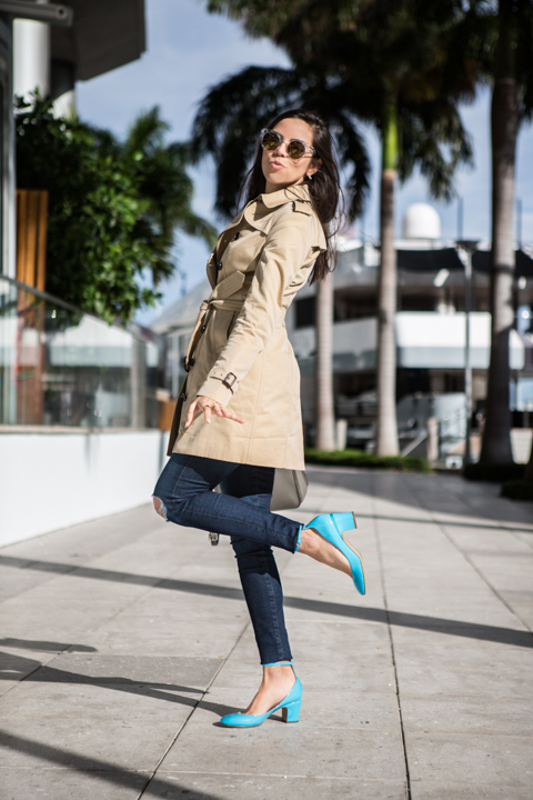 A woman in a pale jacket and bright heels poses on a sunny sidewalk.