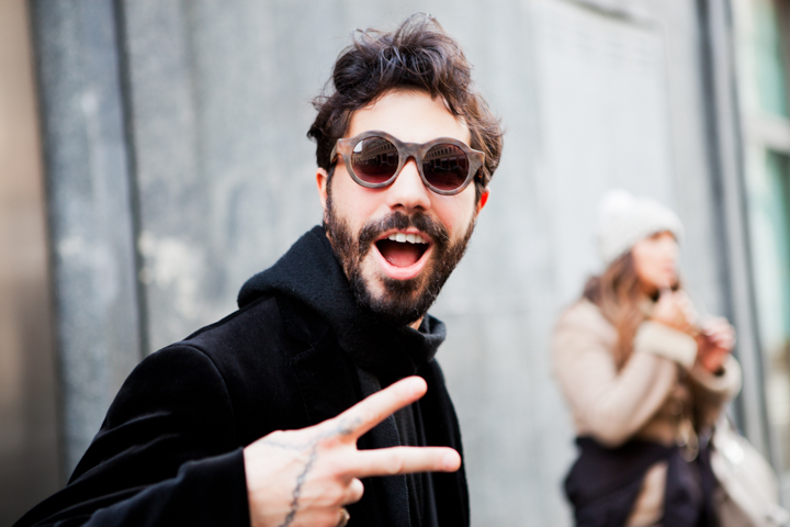 A smiling man in sunglasses raises two fingers on a city street.