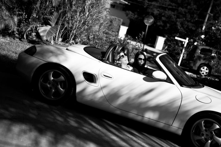 Two adults sit in a convertible beneath trees in black and white.