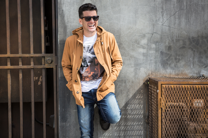 A man in sunglasses and a tan jacket leans against a concrete wall.