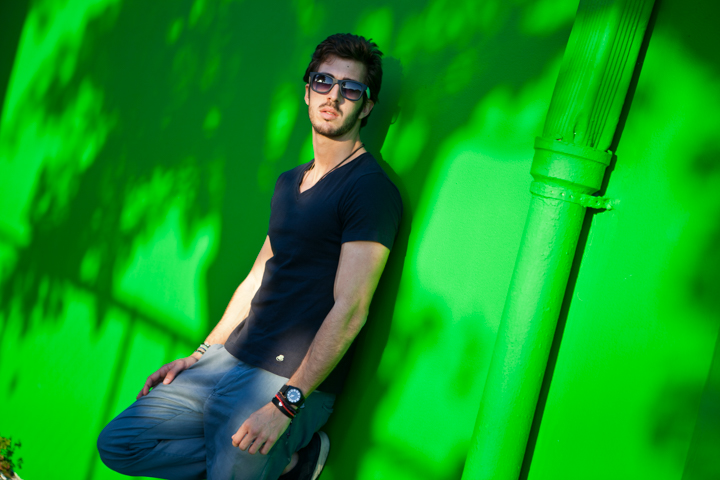 A man wearing sunglasses sits beside a vivid green wall.