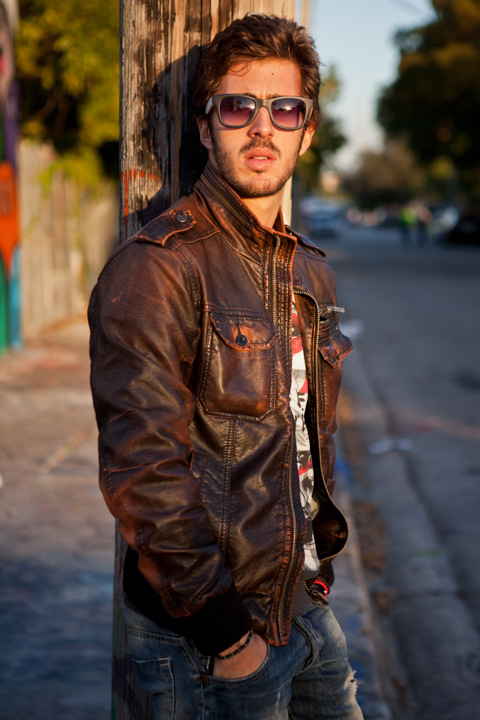 A man in sunglasses and a brown leather jacket on a sunlit street.