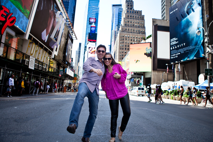 Two adults jump together on a busy city street beneath large billboards.