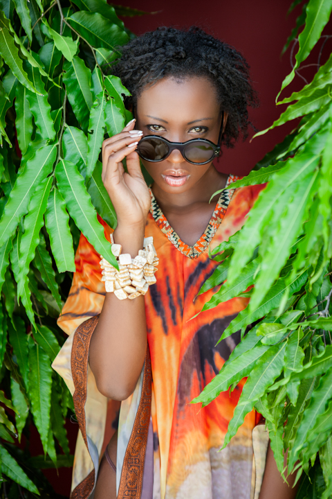 A woman in sunglasses and colorful clothing sits among hanging green leaves.