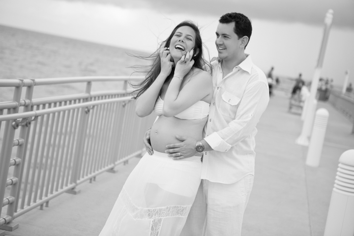 A man embraces a pregnant woman beside a seaside railing in black and white.