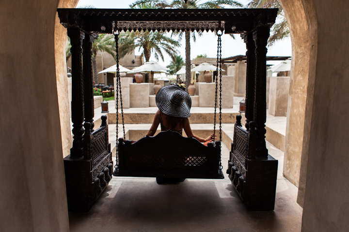 A person in a hat sits on a swing beneath a covered passageway.