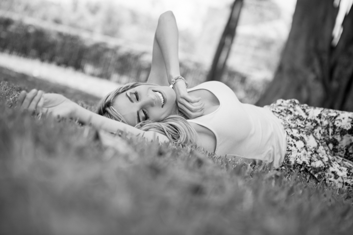 A woman lies on grass beside a tree with her eyes closed.