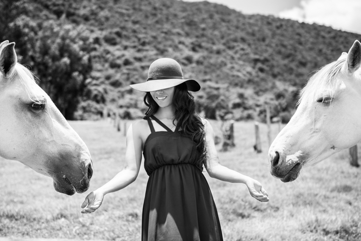 A woman in a wide-brimmed hat stands between two white horses in black and white.