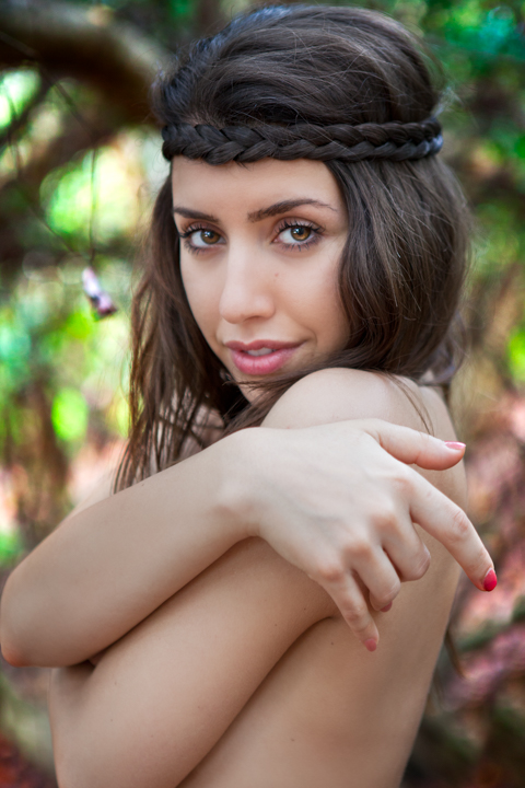 A woman with a braided headband wraps her arms around her shoulders outdoors.