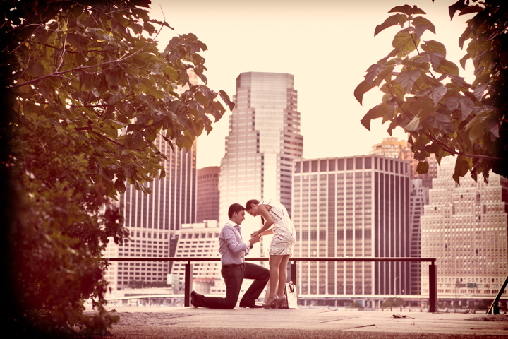 A man kneels in front of a woman beside a waterfront city skyline.