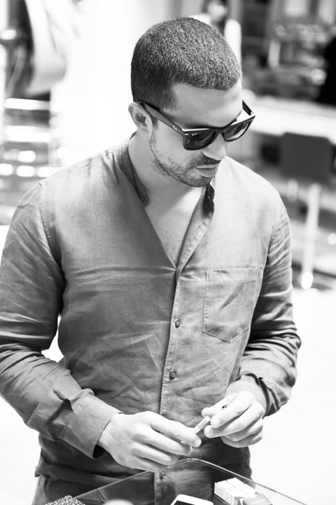 A man wearing sunglasses looks down while buttoning his shirt.