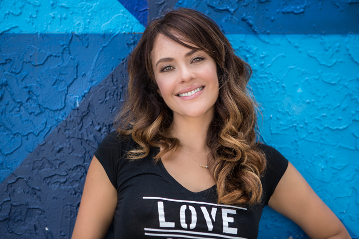 A smiling woman stands against a blue painted wall.