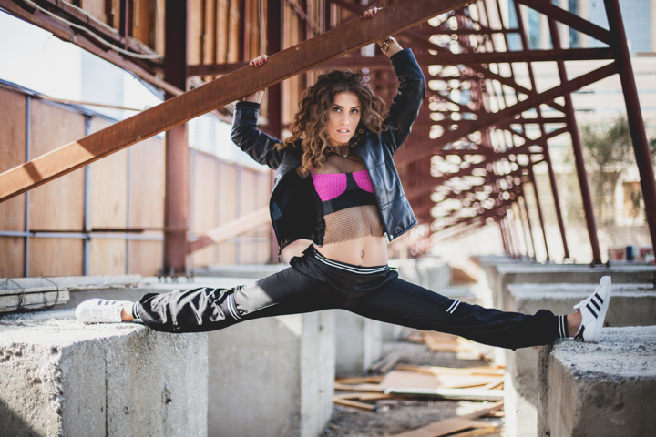 A woman balances in a wide split on a concrete structure beneath metal beams.