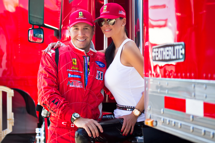 Two adults smile together beside a red racing vehicle.