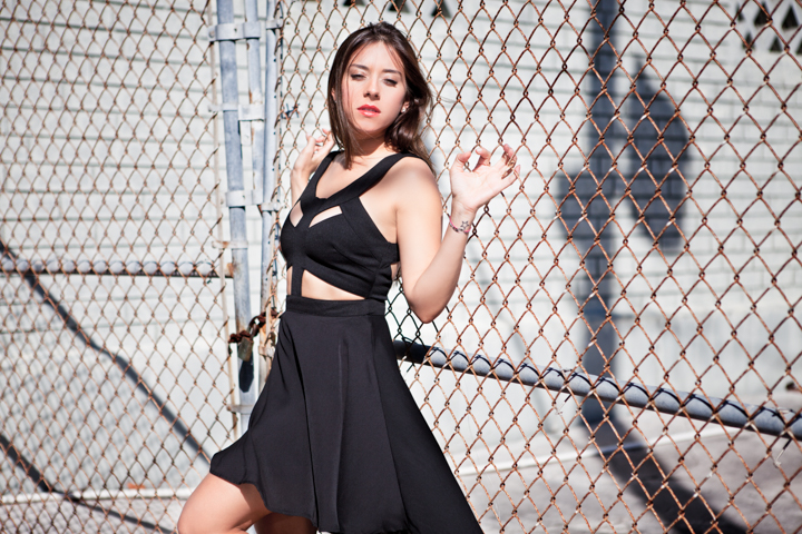 A woman in a black outfit poses beside a chain-link fence.