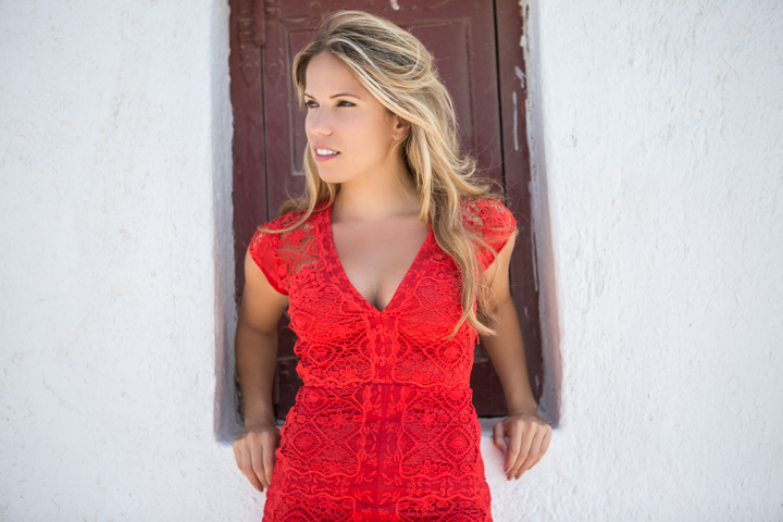 A woman in a red lace dress stands in a dark window frame.