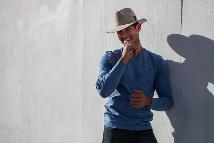 A man in a hat and blue shirt stands against a sunlit white wall.