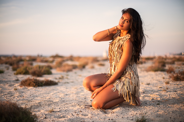 A woman in a patterned dress kneels on rocky sand at sunset.