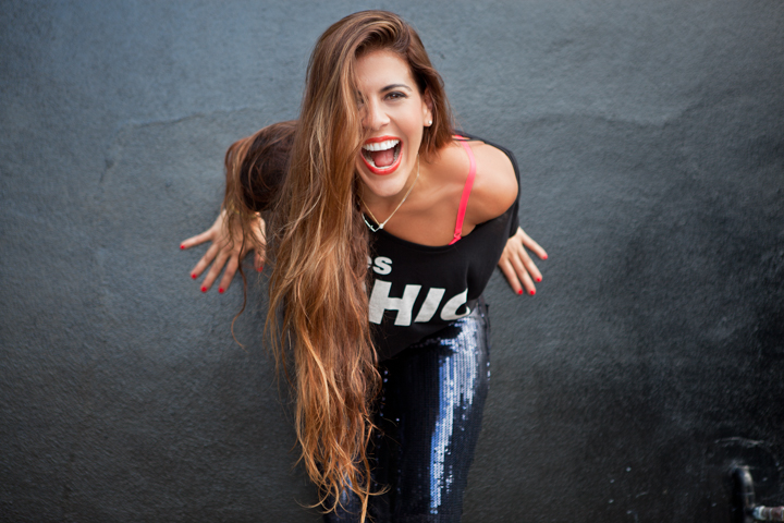 A laughing woman with long hair leans forward against a dark wall.
