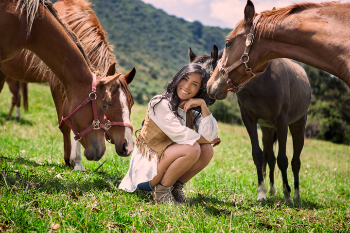A woman crouches in a green pasture beside brown horses.