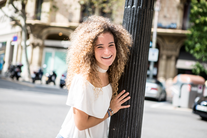 A smiling woman with curly hair rests her hand on a street lamp.