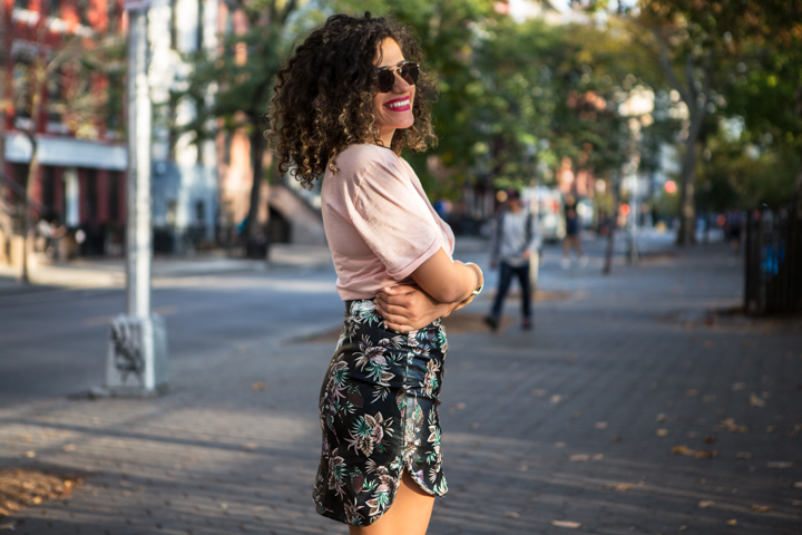 A woman in sunglasses and a printed skirt stands with folded arms on a city street.