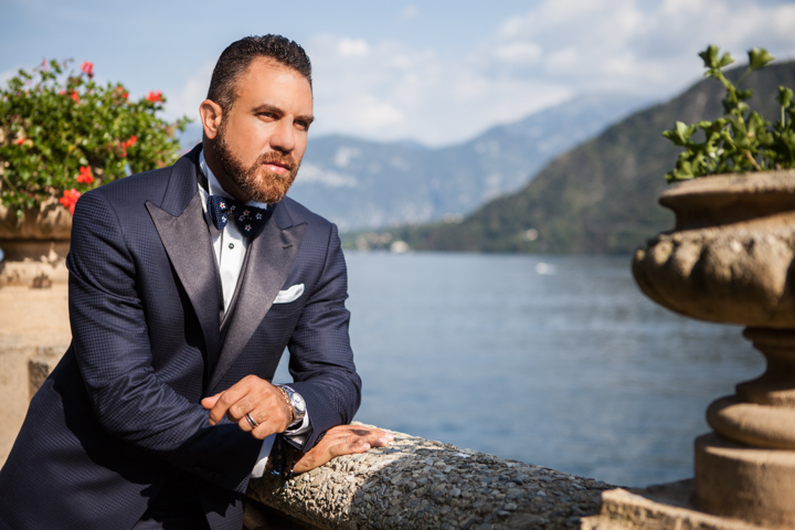 A man in a suit and bow tie leans on a stone railing above a lake with mountains behind.