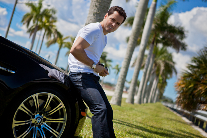 A smiling man holds a drink while leaning beside a dark car.