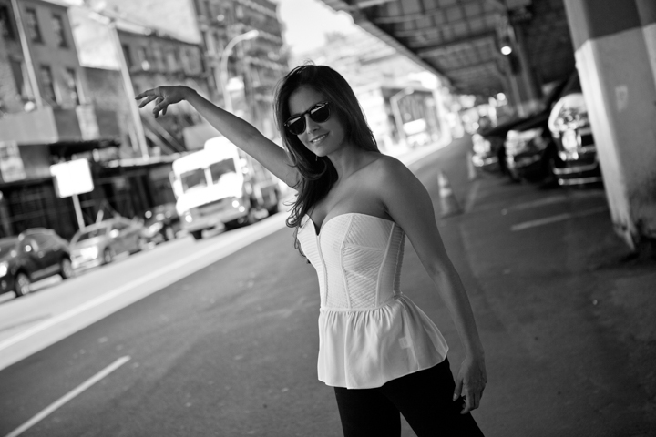 A woman in sunglasses and a white blouse stands on a city street in black and white.