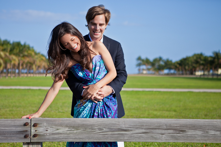 A man hugs a smiling woman from behind beside a park bench.