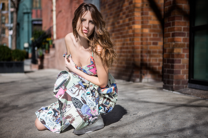 A woman in a colorful dress sits on a sidewalk beside a brick building.