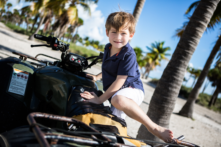 A smiling child sits on an all-terrain vehicle beneath palm trees.