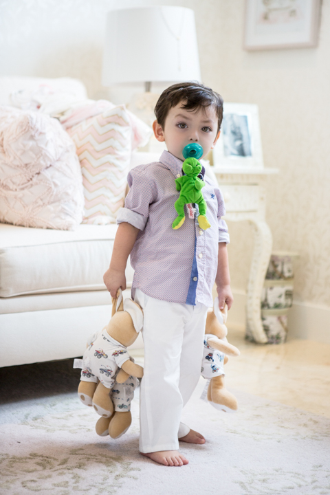 A toddler holds a green toy near their face while standing indoors.