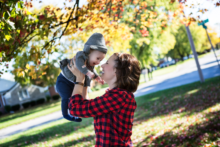 A woman lifts a small child beneath autumn leaves.