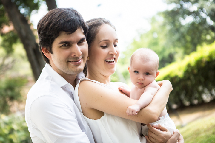Two adults hold a baby close together in a green park.